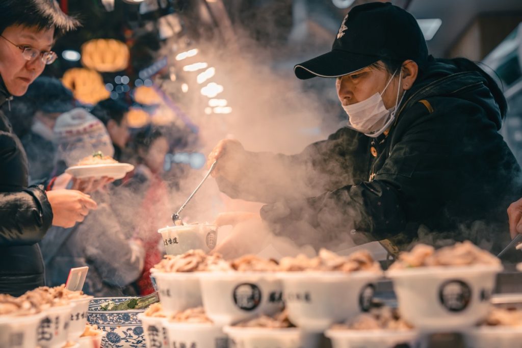 A man serves hot soup at a street trader stall; he wears a mask whilst serving, so is aware of the need for good street food hygiene.