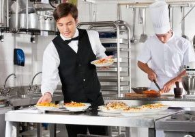 Young waiter placing dishes in tray with chef working in commercial kitchen